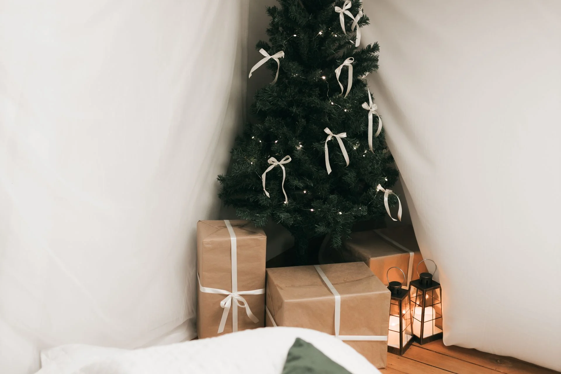 A warm indoor christmas scene with a tree with white ribbon bows, kraft-wrapped gifts, and glowing lanterns